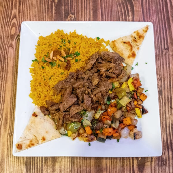 A white plate with yellow rice, sliced meat, roasted vegetables, and pita bread on a wooden table.