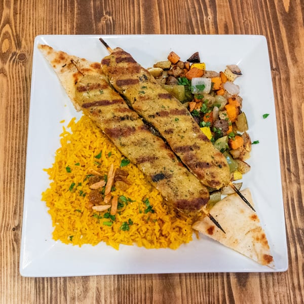 Two grilled chicken skewers with yellow rice, roasted vegetables, and pita bread on a white plate.