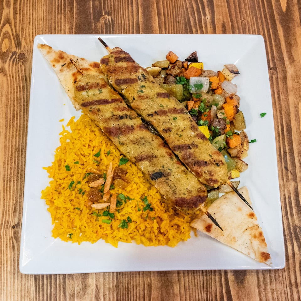 Two grilled chicken skewers with yellow rice, roasted vegetables, and pita bread on a white plate.