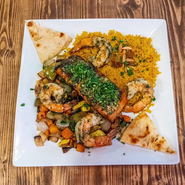 A salmon and shrimp dish with yellow rice, roasted vegetables, and pita bread on a white plate.
