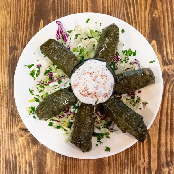 Six stuffed grape leaves arranged around a small bowl of white sauce with red spice on a white plate.