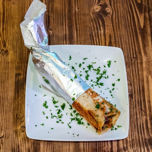 Shawarma wrap, partially unwrapped from foil, on a white plate with scattered parsley on a wooden table.