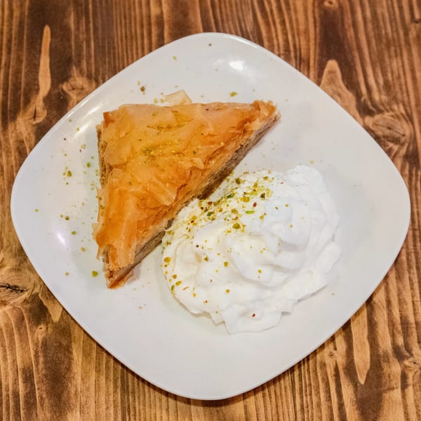 A triangular piece of baklava with whipped cream and pistachio crumbles on a white plate.