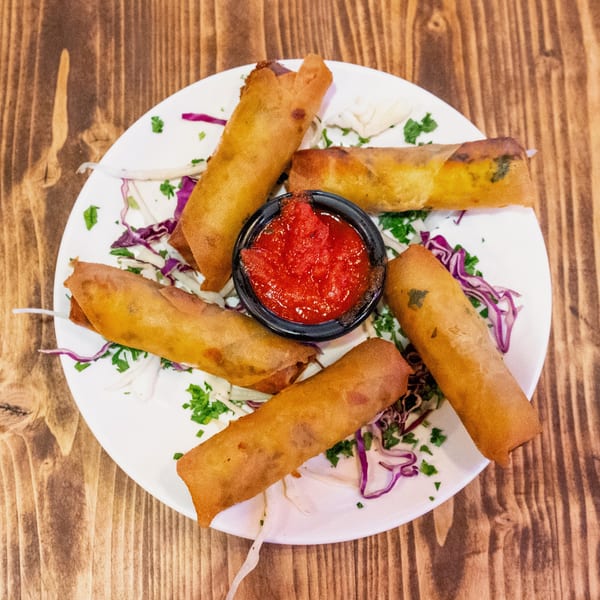 Six crispy spring rolls arranged around a small bowl of red dipping sauce on a white plate.