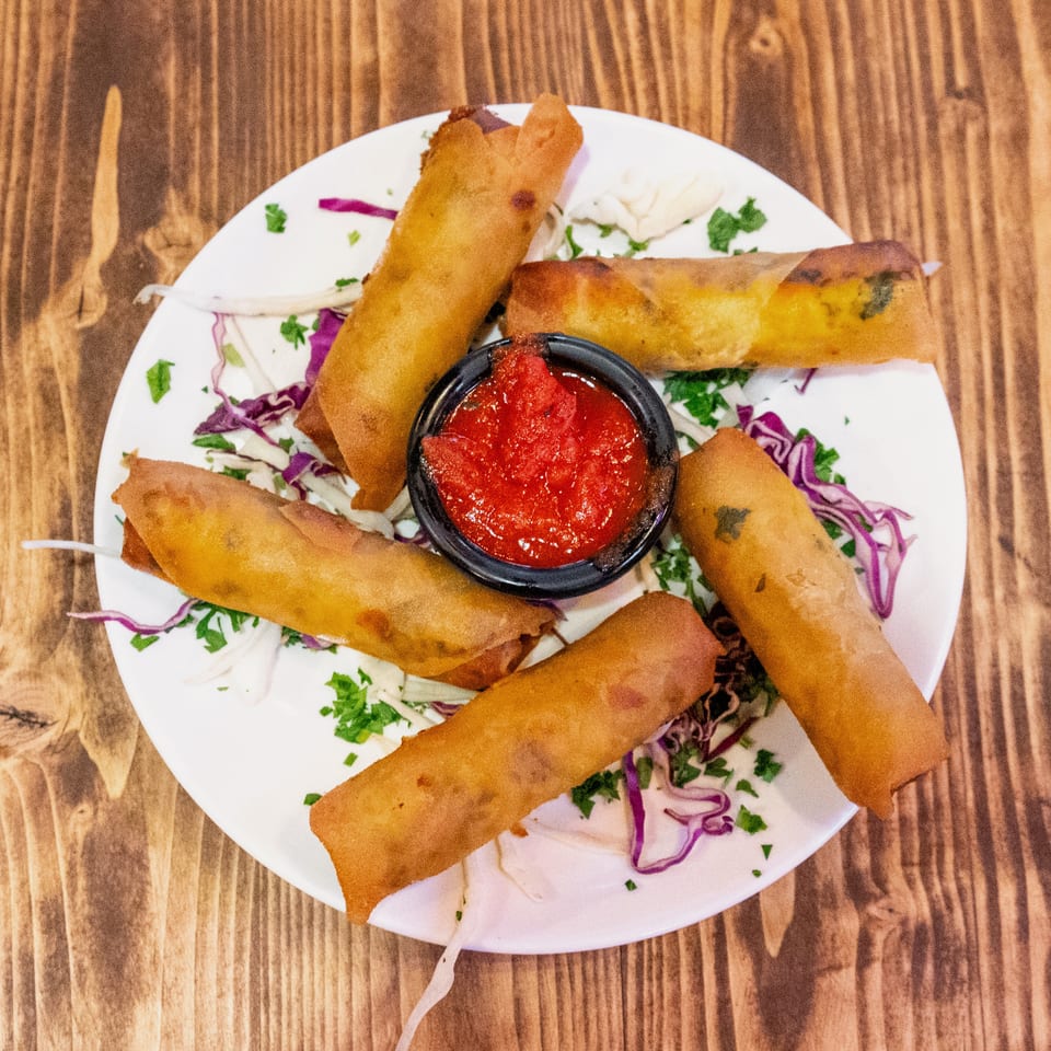 Six crispy spring rolls arranged around a small bowl of red dipping sauce on a white plate.