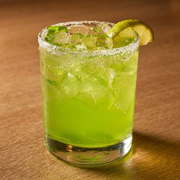 Bright green cocktail with ice, a salted rim, and a lime wedge on a wooden table.