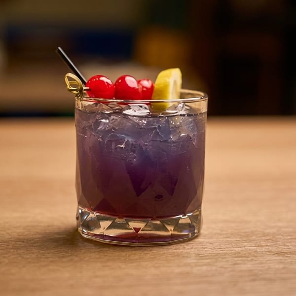 A vibrant purple cocktail with ice, garnished with cherries and a lemon slice, on a wooden table.