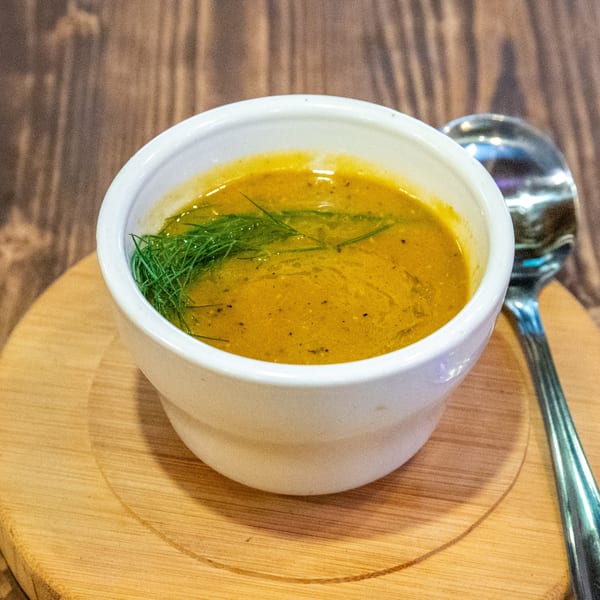 A white bowl of golden soup with green herbs on a wooden board, with a spoon beside it.