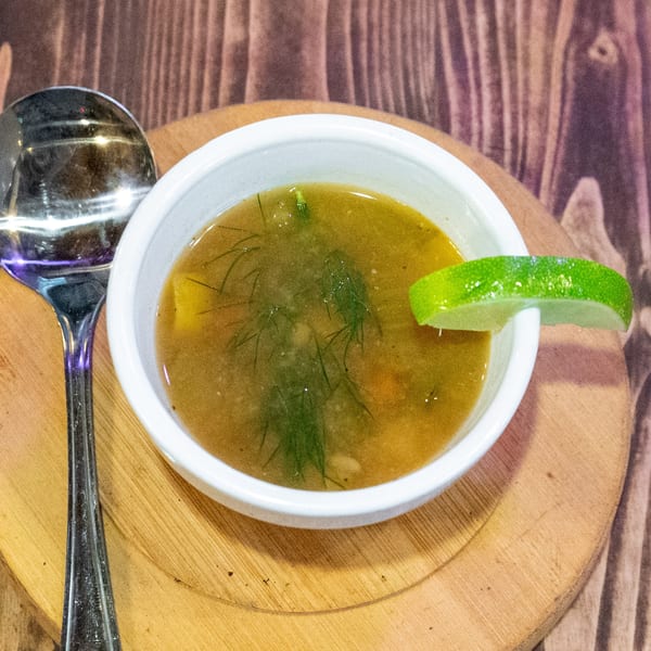 A bowl of soup with dill and a lime wedge, next to a spoon on a wooden board.